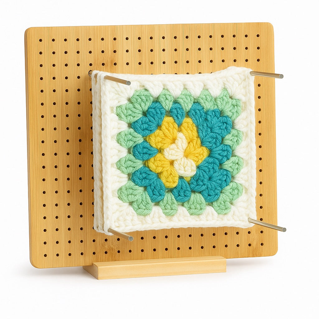 Tablero de bloqueo de madera de 20×20 cm con varillas metálicas y pieza de crochet de colores pastel sujeta en el centro, sobre fondo blanco.