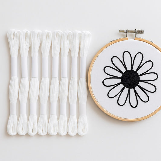 Madejas de hilo Mouliné blanco junto a un bastidor de bordado con diseño minimalista de flor delineada en hilo negro sobre fondo blanco.