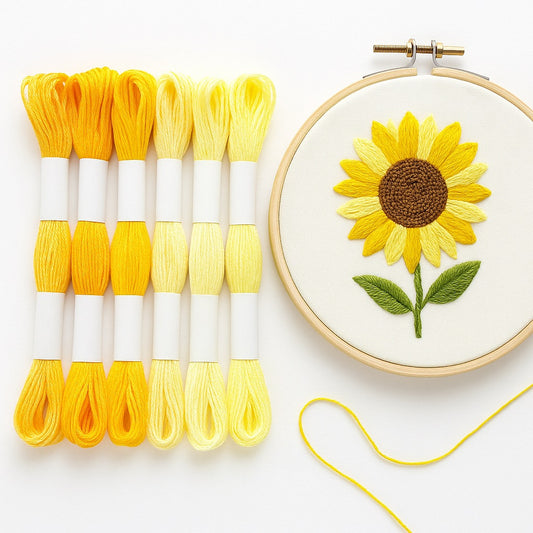 Madejas de hilo Mouliné en tonos amarillos junto a un bastidor con bordado moderno de girasol en degradado de amarillos sobre fondo blanco.