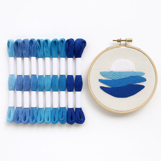 Madejas de hilo Mouliné en tonos azules junto a un bastidor con bordado moderno de diseño marino en degradado de azules sobre fondo blanco.