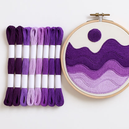 Madejas de hilo Mouliné en tonos morados y lilas junto a un bastidor de bordado moderno con diseño abstracto en degradado de púrpuras sobre fondo blanco.