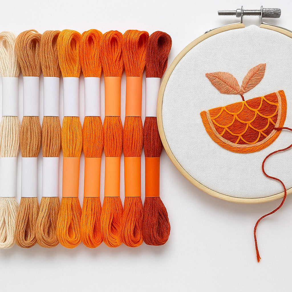 Madejas de hilo Mouliné en tonos naranjas y terracota junto a un bastidor con bordado moderno de una rodaja de naranja con hojas en tonos cálidos sobre fondo blanco.