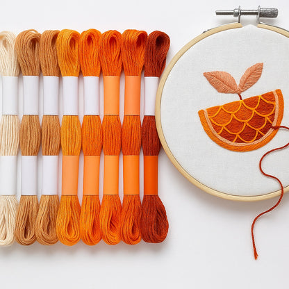 Madejas de hilo Mouliné en tonos naranjas y terracota junto a un bastidor con bordado moderno de una rodaja de naranja con hojas en tonos cálidos sobre fondo blanco.