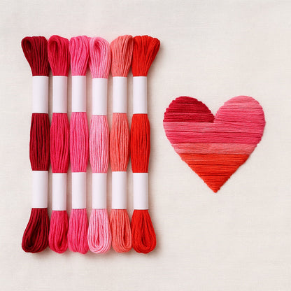 Madejas de hilo Mouliné en tonos rojos y rosados junto a un bordado en forma de corazón degradado en los mismos colores sobre fondo blanco.