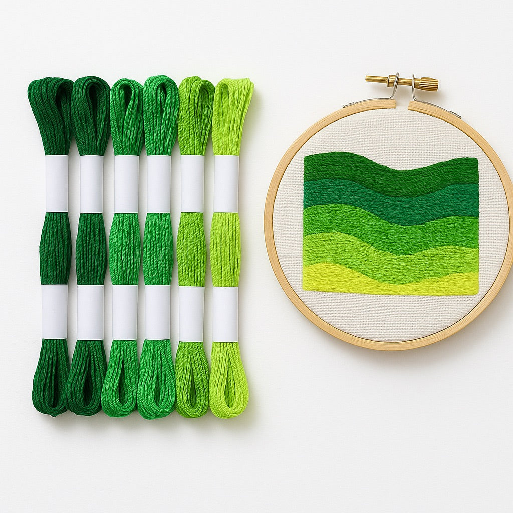 Madejas de hilo Mouliné en tonos verdes junto a un bastidor con bordado moderno de diseño botánico en degradado de verdes sobre fondo blanco.