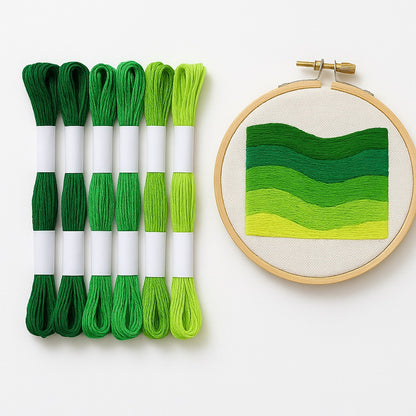 Madejas de hilo Mouliné en tonos verdes junto a un bastidor con bordado moderno de diseño botánico en degradado de verdes sobre fondo blanco.