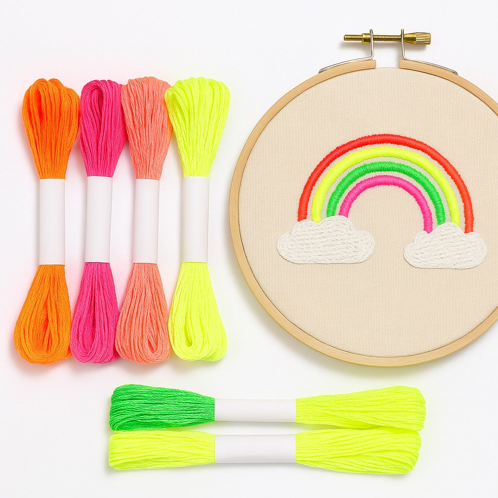 Madejas de hilo Mouliné fluorescente en colores neón junto a un bastidor de bordado con diseño de arcoíris bordado en hilos naranja, rosa, verde y amarillo sobre fondo blanco.