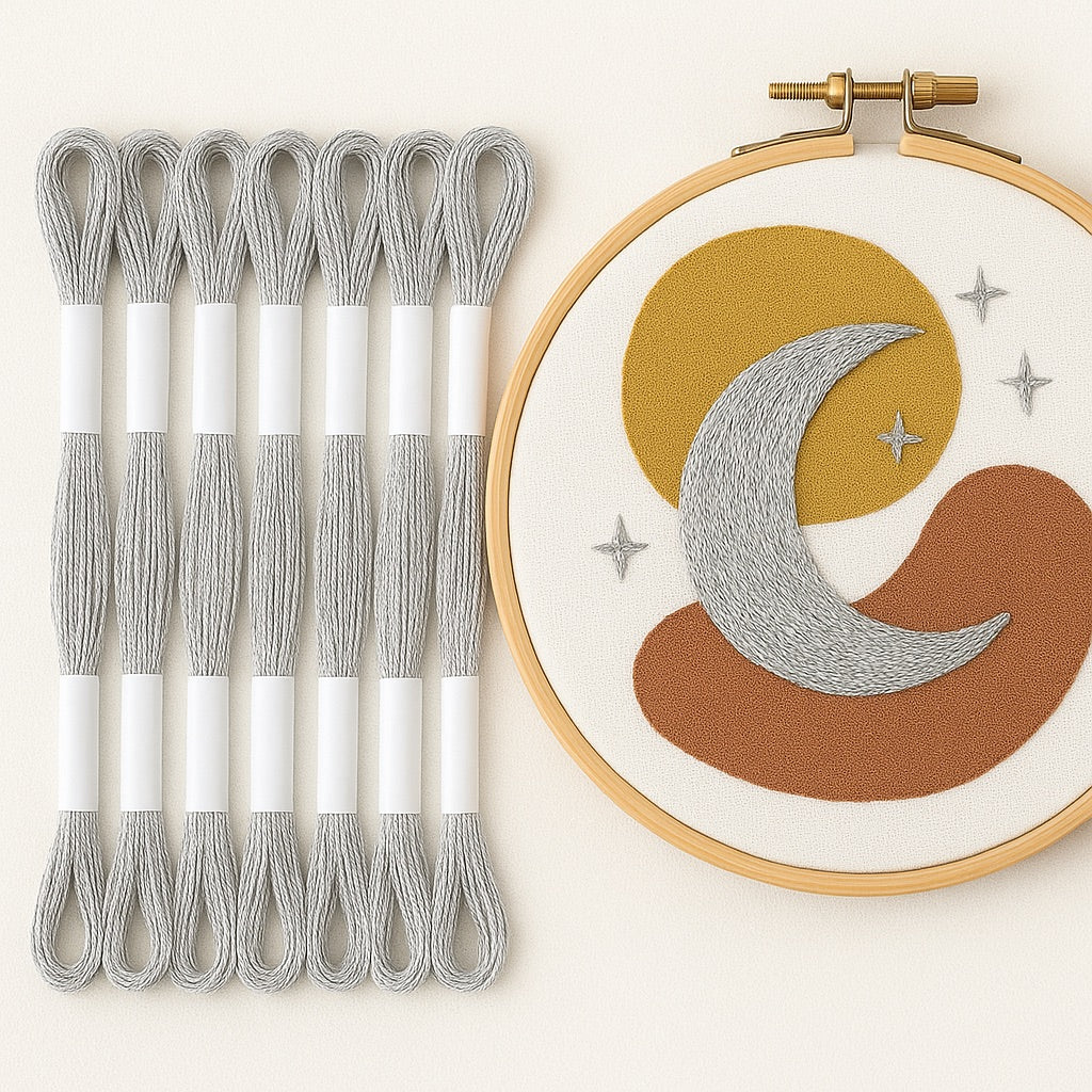 Madejas de hilo Mouliné metalizado color plata junto a un bastidor de bordado moderno con diseño de luna y estrellas bordadas en hilo plateado sobre fondo blanco.