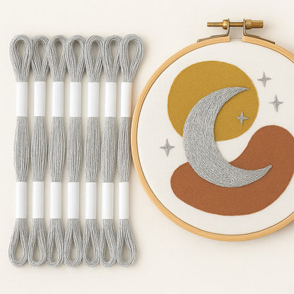 Madejas de hilo Mouliné metalizado color plata junto a un bastidor de bordado moderno con diseño de luna y estrellas bordadas en hilo plateado sobre fondo blanco.