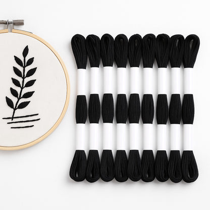 Madejas de hilo Mouliné negro junto a un bastidor con bordado moderno de hoja minimalista en hilo negro sobre tela blanca.